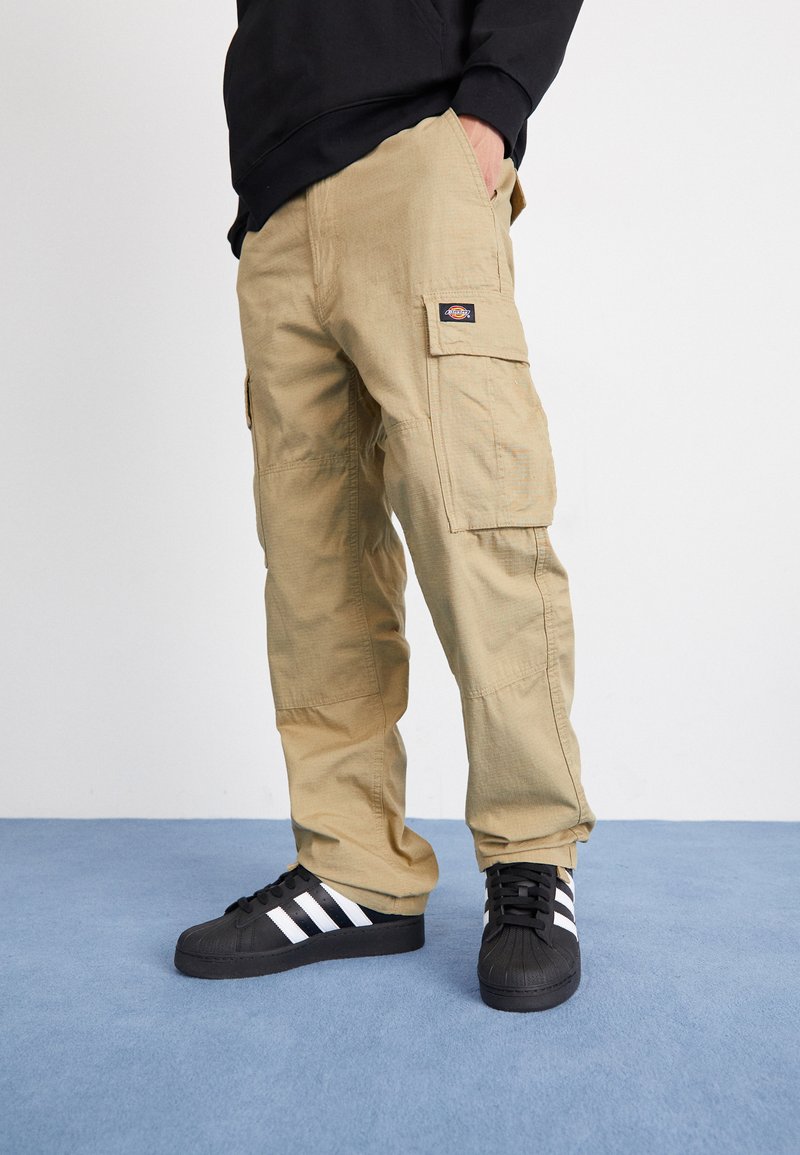Pantaloni cargo beige realizzati in tessuto resistente, caratterizzati da molteplici tasche, cuciture rinforzate e un logo a toppa sulla coscia. Indossati con sneakers nere.