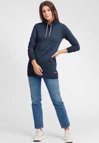 Mörkblå hoodieklänning med cowl-hals, utrustad med dragsnören och en framficka, kombinerad med ljusblå jeans och vita sneakers.