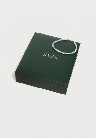 Γυαλιστερό σκούρο πράσινο κουτί με χρυσό κείμενο και στρογγυλό βραχιόλι με χάντρες σε ροζ, μπλε και μοβ χρώματα, τοποθετημένο πάνω του σε λευκό φόντο.