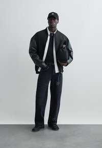 Fekete bomber dzseki szürke bőr ujjal, fehér ing, fekete póló, kopott fekete farmer szegecses részletekkel, és fekete sportcipő.