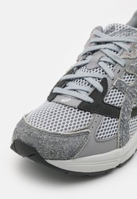 Grå och vit idrottssko med meshöverkropp, mockadetaljer, ljusgrå snören och en svart texturerad sula, visad från framsidan.