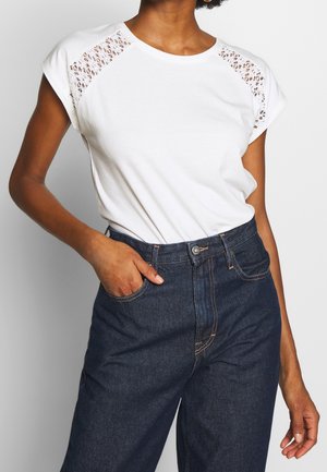 Personne portant un t-shirt blanc orné de dentelle, rentré dans un jean en denim foncé taille haute, avec une main dans une poche.