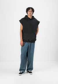 Zwarte mouwloze hoodie met een voorzak en een capuchon met trekkoord, gecombineerd met lichtblauwe wijde jeans en zwarte sneakers.