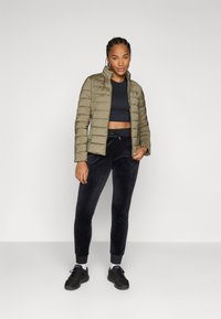 Doudoune vert olive avec un design matelassé, crop top noir et pantalon de survêtement sombre. Des baskets noires avec des accents blancs complètent la tenue.