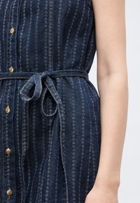 Φόρεμα χωρίς μανίκια από σκούρο μπλε τζιν με κάθετες λευκές ραφές, μπροστινά κουμπιά και ζώνη μέσης δεμένη σε κορδόνι πάνω στον κορμό και το χέρι ενός ατόμου.