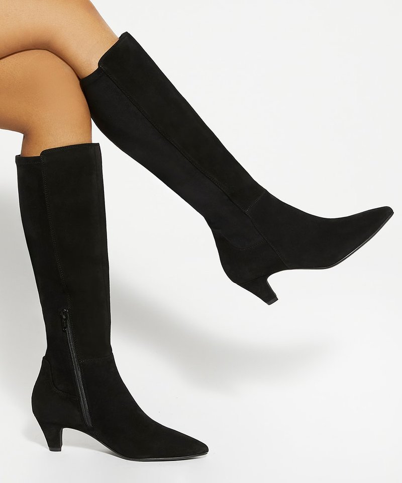 Kruisende benen met zwarte kniehoge suède laarzen met puntige neuzen, zijritsen en lage kitten heels op een witte achtergrond.