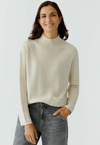 Cremefarbener Pullover mit hohem geripptem Kragen, langen Ärmeln und einer kleinen Fronttasche; kombiniert mit dunkelgrauen Jeans.