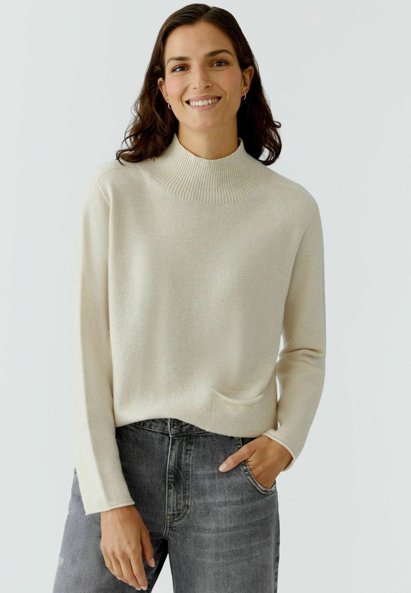 Cremefarbener Pullover mit hohem geripptem Kragen, langen Ärmeln und einer kleinen Fronttasche; kombiniert mit dunkelgrauen Jeans.