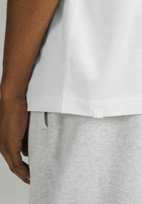 Kortärmad vit t-shirt i bomull med rak nederkant, prydd med en liten vit etikett på sidan, bärs över grå mjukisbyxor.