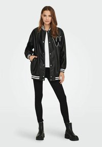 Jachetă varsity din faux piele neagră, cu manșete și guler ribb din material alb cu dungi, având un logo brodat alb cu litera "W" pe piept.