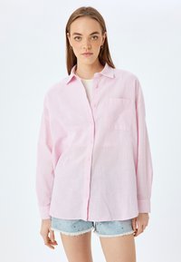 Camisa rosa claro de botones en un ajuste relajado, con un bolsillo en el pecho, mangas largas y una tela texturada parecida al lino.