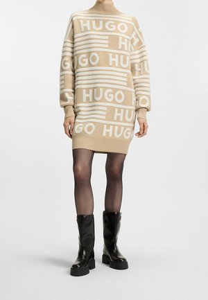 Beige en wit gebreide sweaterjurk met het opschrift "HUGO", gedragen met doorschijnende zwarte panty en zwarte grove mid-kuitlaarzen tegen een effen achtergrond.