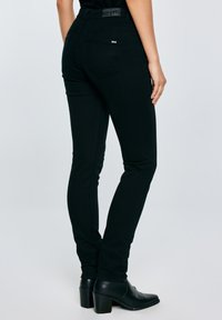 Nő fekete, testhezálló farmernadrágban és fekete, sarkú bokacsizmában, egyik kezét nyugodtan oldalt tartva, világos, egyszerű háttér előtt áll.