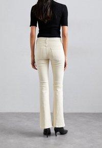 Jean blanc évasé en denim, avec des poches arrière et des détails de couture subtils, associé à des chaussures à talons noirs.