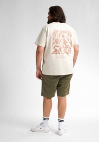 Beige katoenen t-shirt met een koraalvormig bloemenmotief op de achterkant, gecombineerd met olijfgroene shorts. Draag het met witte sneakers en gestreepte sokken.