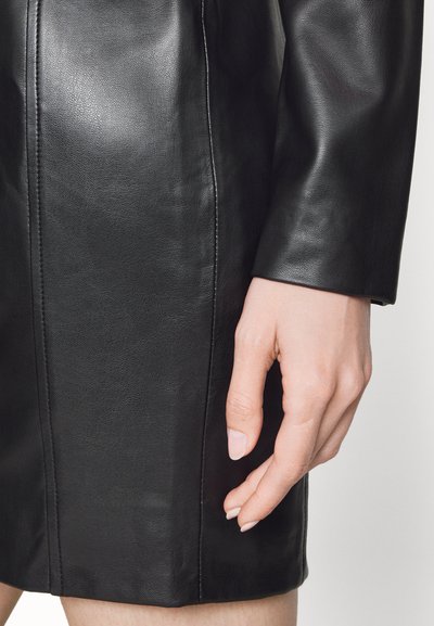 Čierne kožené šaty s hladkou textúrou, s priliehavým dizajnom a viditeľnými švami. Ruka jemne odpočívajúca na látke.