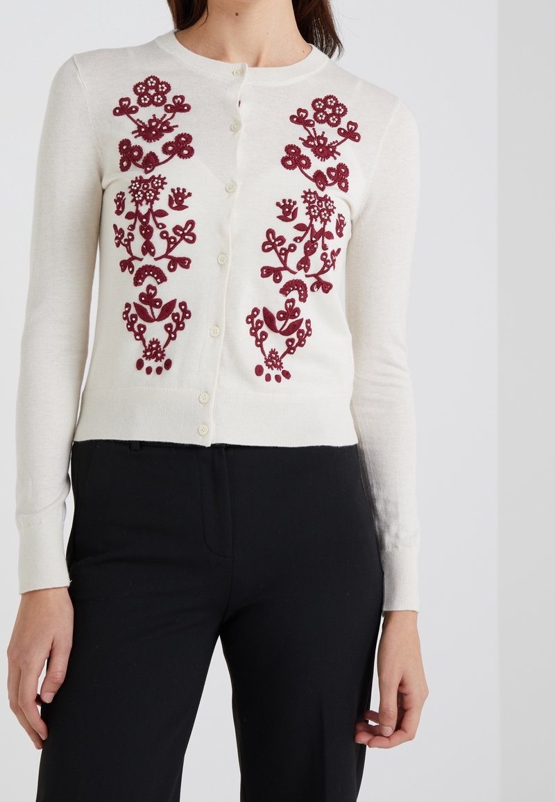 Kreemi kardigan, millel on ees burgundi puna punane lilleline tikand, ümmargune kaelus ja nööbist sulgemine. Pehme kudum, istuv disain.