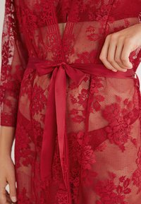 Czerwona koronkowa szlafrok z kwiatowymi wzorami, przezroczystą teksturą, rękawami do połowy przedramienia oraz pasującym satynowym paskiem związanym w talii.
