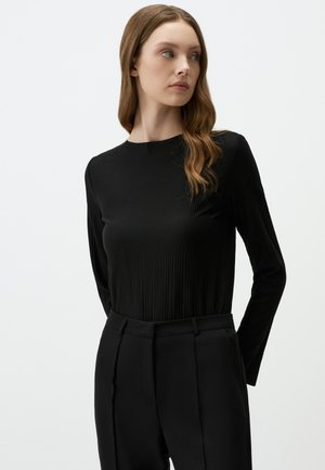 Maglietta nera a maniche lunghe con texture a costine e colletto alto, abbinata a pantaloni neri su misura. Design semplice e lineare.