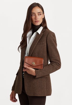 Mujer con cabello largo y castaño lleva una chaqueta de espiga marrón sobre una camisa blanca y un cuello alto negro, sosteniendo una billetera de cuero marrón con logo dorado.