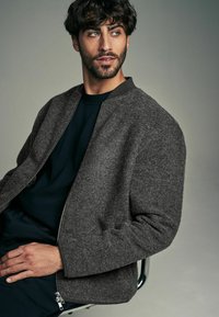 Grijze, gestructureerde bomberjack met een rits, geribbelde kraag en mouwen, gedragen over een donker shirt. Het model zit op een stoel.