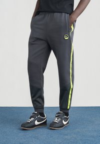 Pantaloni della tuta grigi con accenti neri e strisce laterali giallo neon, caratterizzati da un logo esagonale Nike sulla coscia sinistra e una vita elasticizzata.