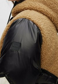 Giacca in shearling marrone con maniche in nylon nero a contrasto, caratterizzata da un patch quadrato. Materiale testurizzato dall'aspetto morbido.