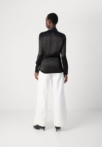 Camicia nera semi-trasparente con maniche lunghe e colletto, abbinata a pantaloni bianchi a gamba larga e tacchi neri. Focus sulla trama liscia e la vestibilità.