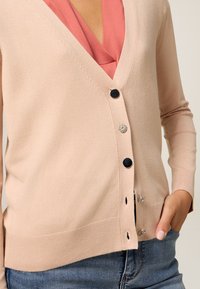 Kvinna som bär en beige cardigan med blandade svarta och silverknappar över en rosa blus, med handen i fickan, och synliga blå jeans.