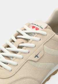 Baskets beige clair avec une texture douce en suède, des lacets blancs et un motif ondulé beige subtil. Dotées d'une étiquette avec logo et d'une semelle en caoutchouc marron.