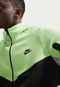Mand iført en neongrøn og sort Nike lynlåstrøje med høj krave, vendt mod venstre mod en ensartet lys baggrund.