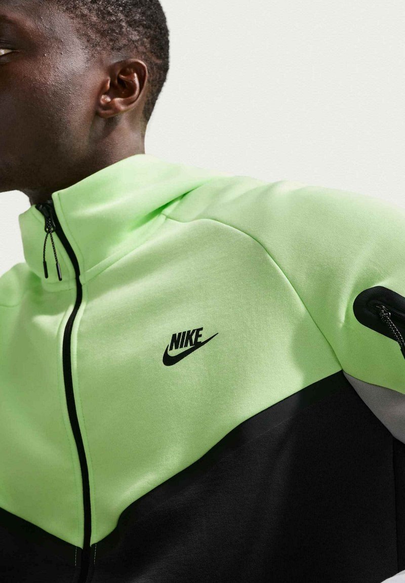 Mand iført en neongrøn og sort Nike lynlåstrøje med høj krave, vendt mod venstre mod en ensartet lys baggrund.