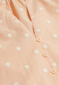 Perzikkleurige blouse met witte geborduurde stippen, voorzien van een horizontale knoopsluiting en een zachte, lichtgewicht stoftextuur.
