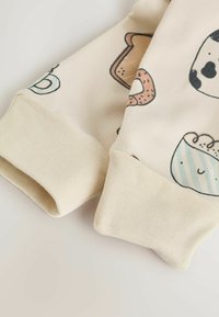 Beżowy materiał z wzorami jedzenia w stylu kreskówkowym, w tym chleb i pączki. Żebrowane mankiety w jednolitym beżowym kolorze. Miękka tekstura, swobodny design.