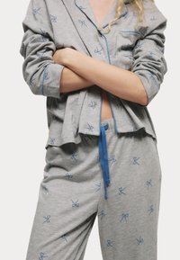 Grå pyjamasset med blåa rosettmönster, bestående av en knappad topp och byxor med snörning. Tillverkat av mjukt, lätt material.