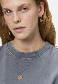 Guldtonet smykke med lagdelte cirkulære vedhæng på en delikat kæde, parret med matchende cirkulære stud øreringe. Båret over en grå sweatshirt.