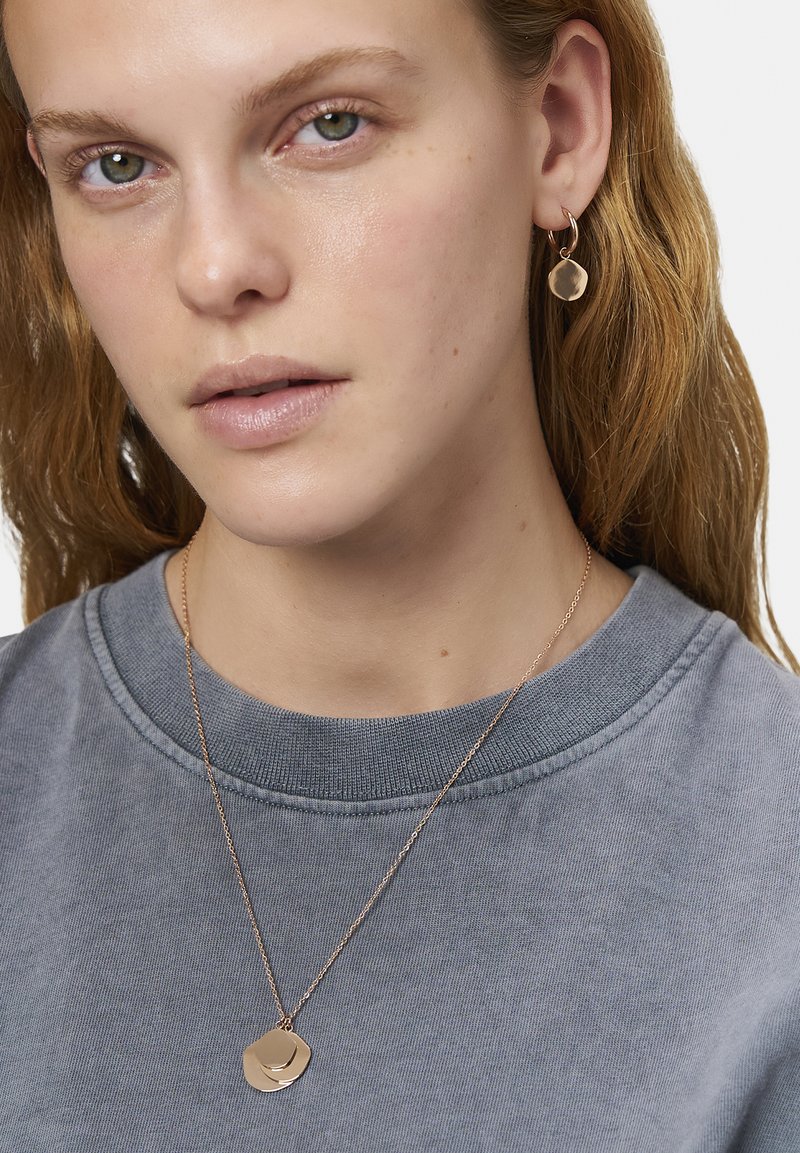 Guldtonet smykke med lagdelte cirkulære vedhæng på en delikat kæde, parret med matchende cirkulære stud øreringe. Båret over en grå sweatshirt.