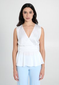 Weißes Satin-Top ohne Ärmel mit V-Ausschnitt und geraffter Taille, mit einem ausgestellten Saum. Kombiniert mit hellblauen, eng anliegenden Hosen.