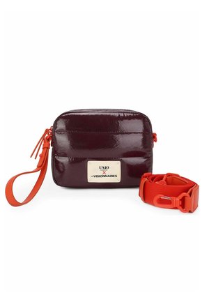 Petit sac rectangulaire violet foncé brillant avec une lanière de poignet orange et une large sangle d'épaule orange amovible. L'étiquette indique « UNIO x LES VISIONNAIRES. »