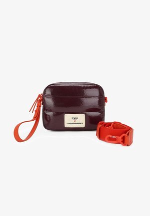 Kleine glanzende donkerpaarse rechthoekige tas met oranje polsbandje en afneembare brede oranje schouderriem. Het label toont "UNIO x LES VISIONNAIRES."