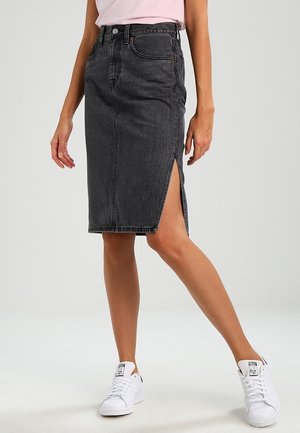 Femme portant une jupe en denim noire taille haute avec fente latérale, un haut rose clair et des baskets blanches, debout devant un fond uni.