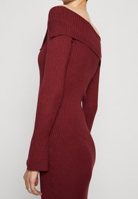 Abito a coste, scollato sulla spalla in bordeaux, design aderente che accentua la silhouette, maniche lunghe e texture sottile. Nessun motivo o dettaglio particolare.