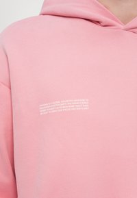 Rosa hettegenser med en teksturert overflate, med hvit trykt tekst på forsiden som beskriver merkenes oppdrag og verdier.