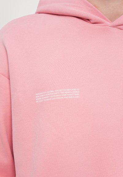 Haut à capuche rose avec une finition texturée, arborant un texte imprimé blanc à l'avant qui décrit la mission et les valeurs de la marque.