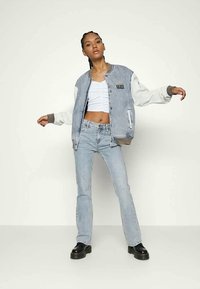 Casaco de ganga com mangas de pele branca em contraste, corpo em ganga azul, bainha canelada e detalhe do logótipos. Combinado com jeans azuis claros de boca larga e sapatos pretos.