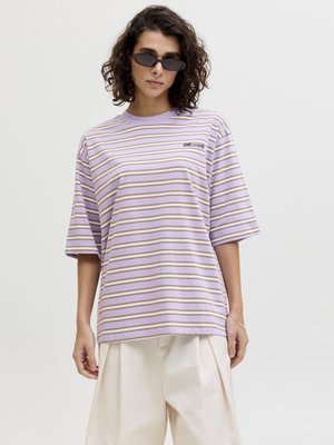 Person med krøllete hår som har på seg svarte, smale solbriller, en oversize T-skjorte med lavendelfargede og beige striper, og løse, hvite bukser.