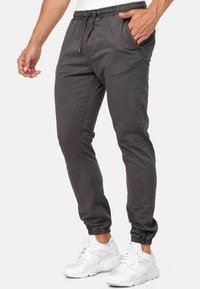 Uomo che indossa pantaloni da jogging grigio scuro con polsini elastici e sneakers bianche, con una mano parzialmente nella tasca, su sfondo semplice.