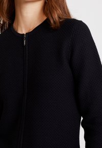 Schwarze Reißverschlussjacke aus strukturiertem Stoff. Verfügt über einen Rundhalsausschnitt und gerippte Bündchen. Schlichtes Design ohne sichtbare Muster oder Akzente.