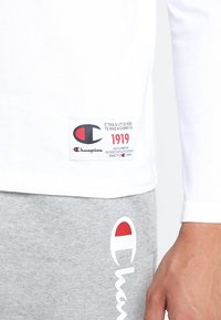 Biała koszula z długim rękawem z metką z tkaniny, na której znajduje się czerwono-czarny logo; zestawiona z szarymi spodniami dresowymi, które mają białe logo z czerwonym akcentem.