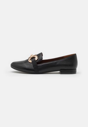 Chaussure plate en cuir noir avec un embout arrondi, ornée d'une boucle circulaire dorée et d'un petit talon carré. Texture lisse avec un design minimaliste.